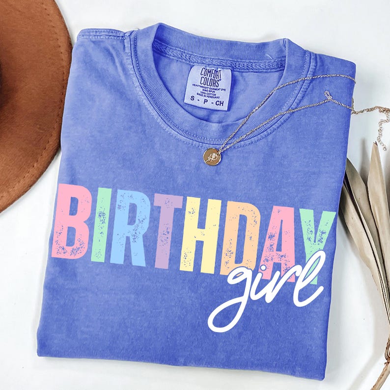 Może przedstawiać: Niebieska koszulka Comfort Colors z napisem "BIRTHDAY girl" w kolorowych literach blokowych. Złoty naszyjnik z małą zawieszką spoczywa na koszulce. Koszulka w rozmiarze S-P-CH.