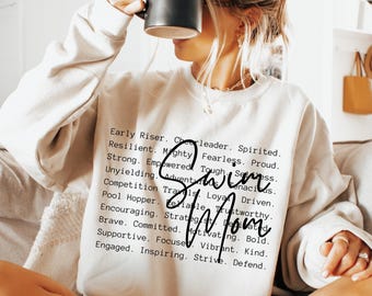 May include: A cream-colored crewneck sweatshirt featuring black text that lists various descriptive words like Early Riser, Resilient, and Strong, with the words Swim Mom written in a large, black, cursive script overlaying the printed text.