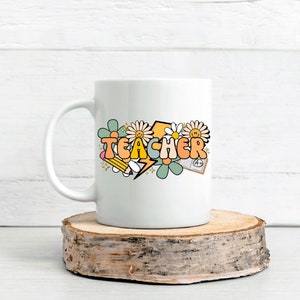 May include: White ceramic mug with a colorful graphic of the word "TEACHER" in a retro font, surrounded by flowers, a pencil, a lightning bolt, and a book with an A+ grade.