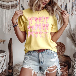 Puede incluir: Una camiseta amarilla con un dise&ntilde;o de lazos rosas. Los lazos var&iacute;an en estilo, algunos con un patr&oacute;n de cuadros. La modelo lleva pantalones cortos vaqueros y un cintur&oacute;n marr&oacute;n. El fondo incluye elementos decorativos.