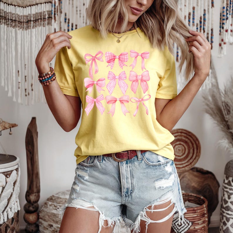 Puede incluir: Una camiseta amarilla con un dise&ntilde;o de lazos rosas. Los lazos var&iacute;an en estilo, algunos con un patr&oacute;n de cuadros. La modelo lleva pantalones cortos vaqueros y un cintur&oacute;n marr&oacute;n. El fondo incluye elementos decorativos.