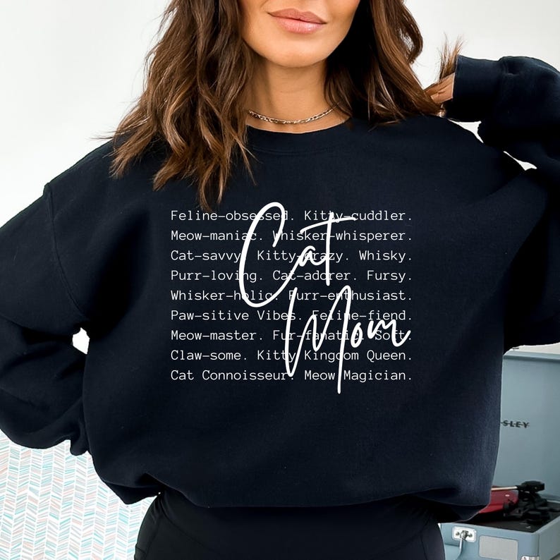 May include: Black sweatshirt with the words "Cat Mom" in a cursive font. The shirt has a list of cat-related words in a white font, including "Feline-obsessed," "Meow-maniac," and "Purr-loving."