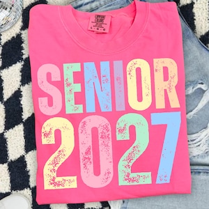 May include: A bright pink t-shirt featuring the text SENIOR 2027 in large, distressed, multi-colored block letters. The shirt has a crew neckline and a Comfort Colors brand label on the inside collar indicating a size large.