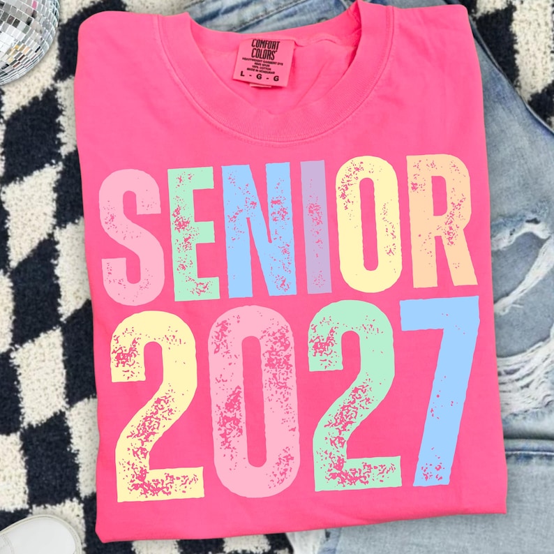 May include: A bright pink t-shirt featuring the text SENIOR 2027 in large, distressed, multi-colored block letters. The shirt has a crew neckline and a Comfort Colors brand label on the inside collar indicating a size large.