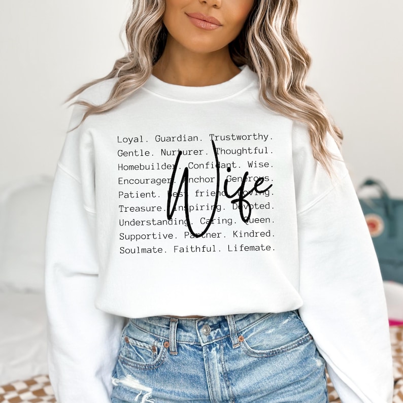 Pu&ograve; includere: Felpa bianca con la parola "Wife" in grandi lettere nere e un elenco di aggettivi positivi che descrivono una moglie in lettere nere pi&ugrave; piccole.
