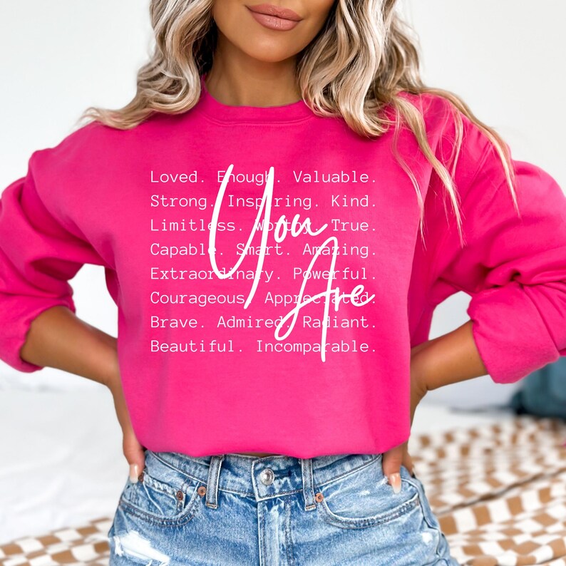May include: Pink sweatshirt with white text that reads "You Are" and a list of positive words describing a person, including loved, enough, valuable, strong, inspiring, kind, limitless, true, capable, smart, amazing, extraordinary, powerful, courageous, appreciated, brave, admired, radiant, beautiful, and incomparable.