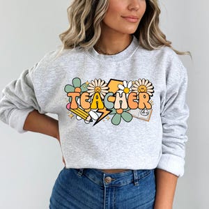 May include: A gray sweatshirt with a graphic design of the word "TEACHER" in colorful, retro-style letters. The design includes flowers, a lightning bolt, a pencil, and a notebook with an A+ grade.