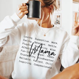 Könnte beinhalten: Ein weißes Langarm-Sweatshirt mit schwarzem Textaufdruck, der verschiedene positive Eigenschaften auflistet, darunter Loving, Supportive, Warm, Caring, Generous, Gentle, Playful, Nurturing, Wise, Affectionate, Fun-loving, Loyal, Happy, Trustworthy, Kindhearted, Reliable, Patient, Chill, Inspiring, Compassionate, mit dem Wort Mama in einer großen schwarzen Schreibschrift in der Mitte.