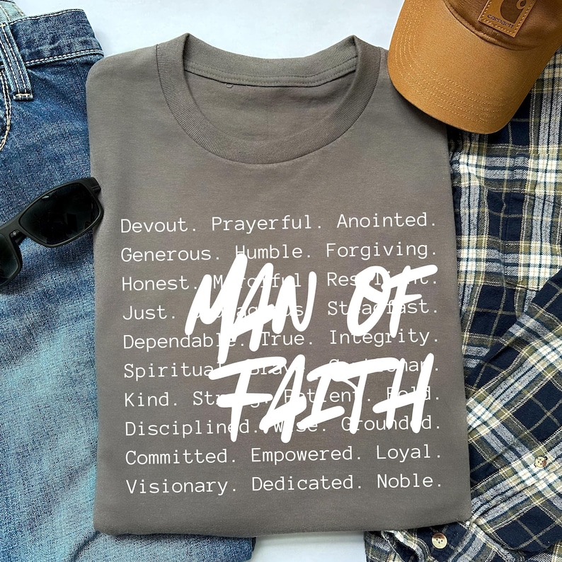 Op de afbeelding: Een grijs t-shirt met de woorden "MAN OF FAITH" in grote witte letters. Het shirt bevat ook een lijst met positieve eigenschappen. Zonnebril, een bruine pet en een geruit overhemd staan ook op de foto.