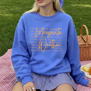 May include: A blue sweatshirt with the words "Margarita Weather" in orange script. The sweatshirt has a crew neck and long sleeves. The text includes words like "Tequila," "Glowing," and "Tropical."