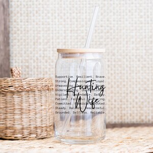 Op de afbeelding: Een doorzichtige glazen beker met een bamboe deksel en een rietje. De beker heeft de woorden "Hunting Wife" in een scriptlettertype, samen met een lijst met ondersteunende kwaliteiten. Een gevlochten mand staat op de achtergrond.