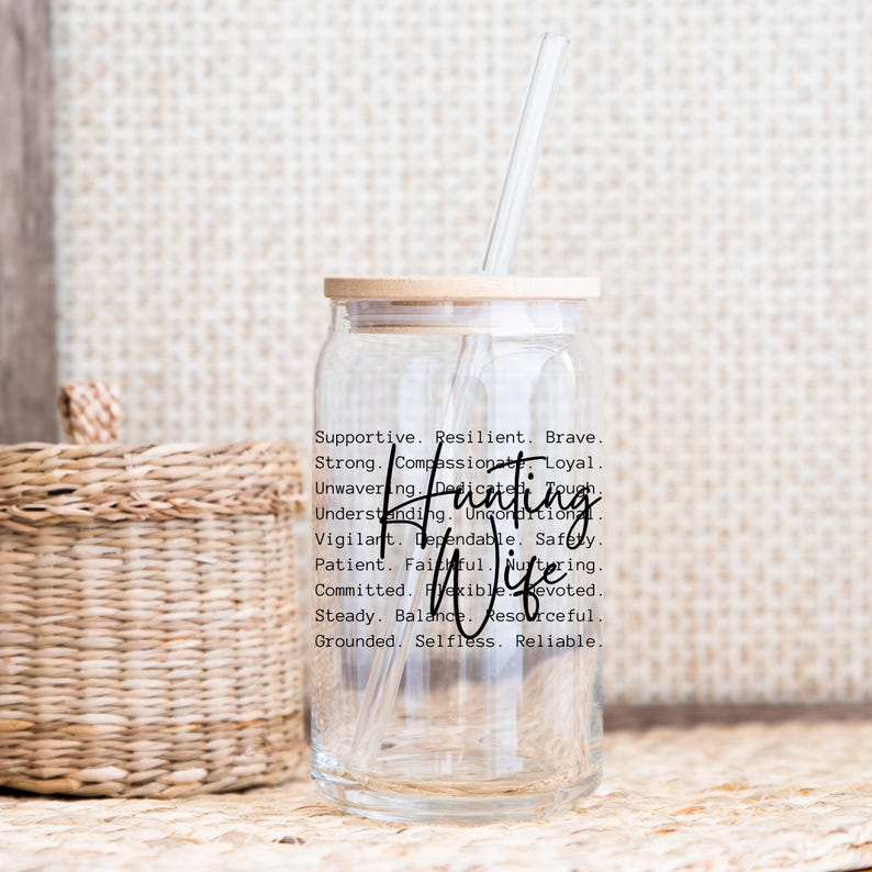Op de afbeelding: Een doorzichtige glazen beker met een bamboe deksel en een rietje. De beker heeft de woorden "Hunting Wife" in een scriptlettertype, samen met een lijst met ondersteunende kwaliteiten. Een gevlochten mand staat op de achtergrond.