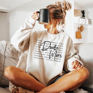 May include: A white sweatshirt with the text "Doodle Mom" in black lettering. The text is surrounded by a list of positive adjectives describing a doodle dog.
