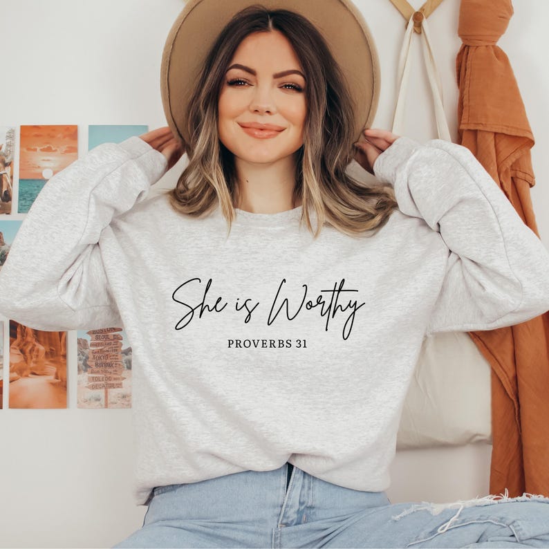 May include: A light grey sweatshirt featuring the script "She is Worthy" and "Proverbs 31" in black. The long-sleeved sweatshirt has a crew neck. The person in the image is wearing a tan hat and blue jeans.