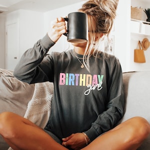 Może przedstawiać: Ciemnoszara koszulka z długim rękawem z napisem "BIRTHDAY girl" w kolorowych pastelowych literach. Koszulka jest wykonana z miękkiego materiału. Czarny kubek jest trzymany przed twarzą.