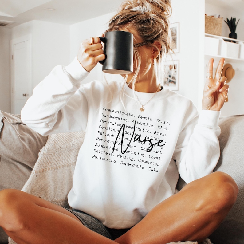 K&ouml;nnte beinhalten: Wei&szlig;er Pullover mit dem Wort "Nurse" in schwarzer Kursivschrift und einer Liste positiver Attribute, die den Beruf beschreiben. Der Text lautet: Mitf&uuml;hlend, Sanft, Klug, Flei&szlig;ig, Aufmerksam, Freundlich, Engagiert, Empathisch, Mutig, Belastbar, Geschickt, Zuverl&auml;ssig, Geduldig, Vertrauensvoll, Beobachtungsgabe, Einfallsreich, Held, Loyal, Unterst&uuml;tzend, F&uuml;rsorglich, Selbstlos, Heilend, Engagiert, Beruhigend, Zuverl&auml;ssig, Ruhig.