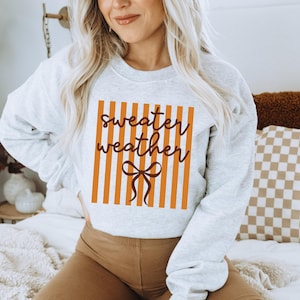 May include: A light gray sweatshirt with the words "sweater weather" in a brown script font, set against a background of orange vertical stripes. A brown bow graphic is below the text. The sweatshirt is a casual, comfortable style.