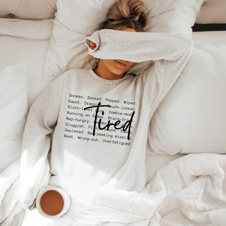 May include: A gray sweatshirt with the word "Tired" printed in black. The text is surrounded by a list of words describing different states of exhaustion.