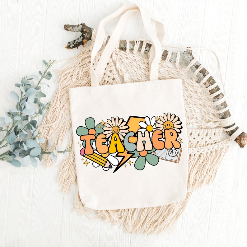 May include: A white canvas tote bag with a colorful graphic design featuring the word "TEACHER" in a retro font, surrounded by flowers, a lightning bolt, a pencil, and a book with an A+ grade.