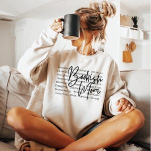 Puede incluir: Una sudadera blanca con el texto "Bookish Mom" en letras negras. El texto est&aacute; rodeado de una lista de palabras que describen a una persona que le gusta leer, como "literaria", "con conocimientos", "perspicaz", "imaginativa", "reflexiva", "amable", "de apoyo", "cordial", "po&eacute;tica", "atractiva", "reconfortante", "alentadora", "consciente", "nost&aacute;lgica", "inspiradora", "sabia", "amante de la fantas&iacute;a".