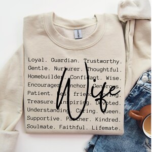 Pu&ograve; includere: Una felpa beige con la parola "Wife" in grandi lettere nere circondata da un elenco di aggettivi positivi che descrivono una moglie. Le parole includono: Leale, Guardiano, degno di fiducia, Gentile, Nutricente, Pensieroso, Costruttore di case, Confidente, Saggio, Incoraggiatore, Ancora, Generoso, Paziente, Miglior amico, Amorevole, Tesoro, Ispiratore, Devoto, Comprensivo, Affettuoso, Regina, Supportivo, Partner, Affine, Anima gemella, Fedele, Compagno di vita.