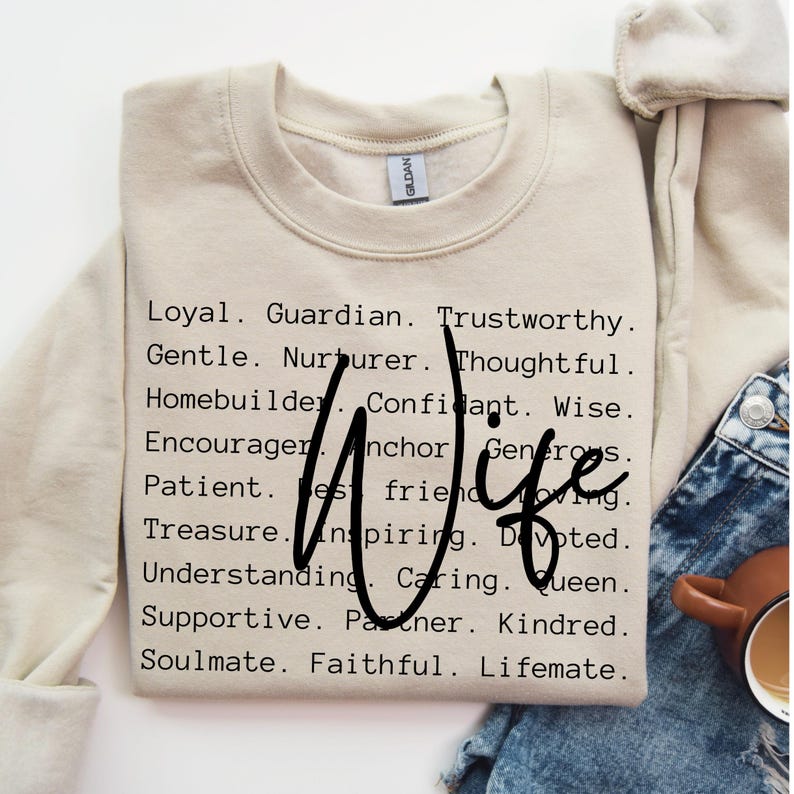 Pu&ograve; includere: Una felpa beige con la parola "Wife" in grandi lettere nere circondata da un elenco di aggettivi positivi che descrivono una moglie. Le parole includono: Leale, Guardiano, degno di fiducia, Gentile, Nutricente, Pensieroso, Costruttore di case, Confidente, Saggio, Incoraggiatore, Ancora, Generoso, Paziente, Miglior amico, Amorevole, Tesoro, Ispiratore, Devoto, Comprensivo, Affettuoso, Regina, Supportivo, Partner, Affine, Anima gemella, Fedele, Compagno di vita.