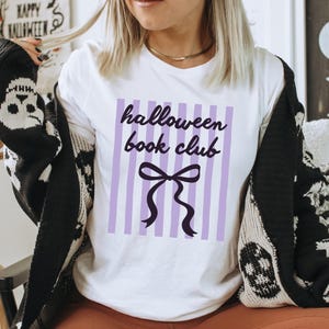 May include: White t-shirt with the text "halloween book club" in black script over a purple striped background. A black bow is below the text. The person is wearing a black and white patterned cardigan.