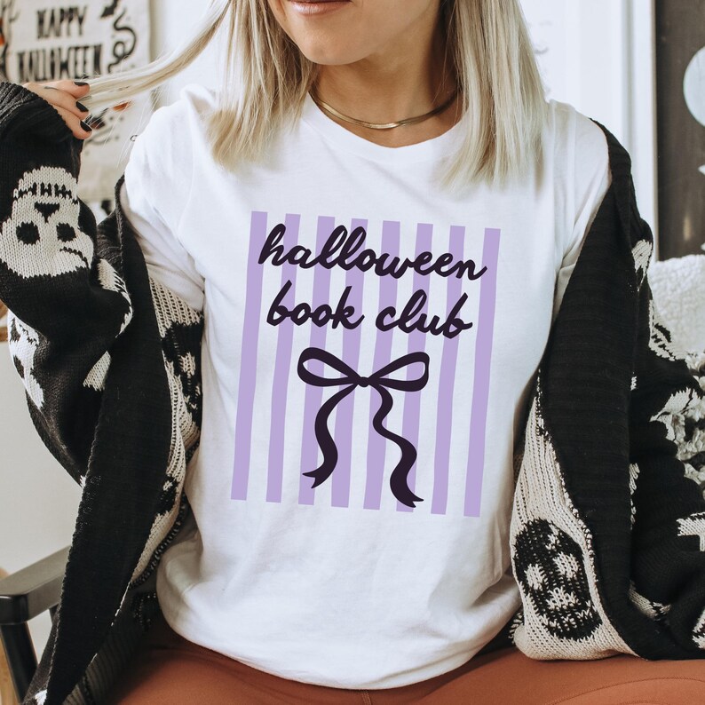 May include: White t-shirt with the text "halloween book club" in black script over a purple striped background. A black bow is below the text. The person is wearing a black and white patterned cardigan.
