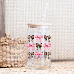 Puede incluir: Un vaso de vidrio transparente con tapa de bamb&uacute; y pajita de vidrio, que presenta un patr&oacute;n repetitivo de lazos rosas y lazos con estampado de leopardo dispuestos en una cuadr&iacute;cula. El dise&ntilde;o est&aacute; impreso en el exterior del vaso cil&iacute;ndrico.