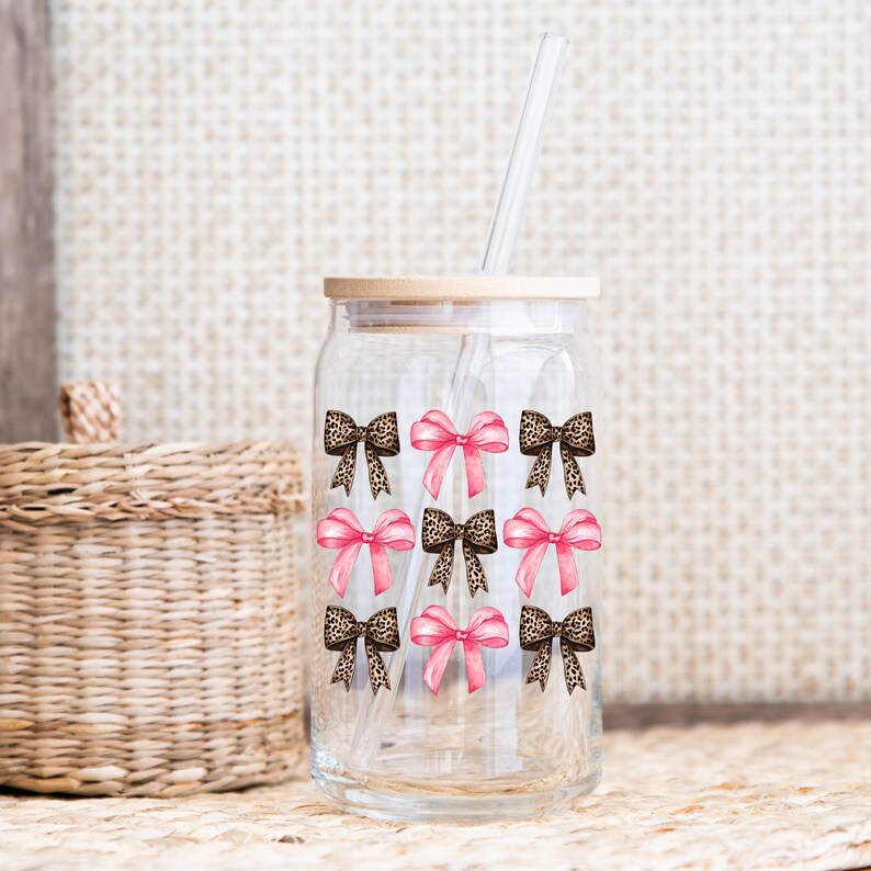 Puede incluir: Un vaso de vidrio transparente con tapa de bamb&uacute; y pajita de vidrio, que presenta un patr&oacute;n repetitivo de lazos rosas y lazos con estampado de leopardo dispuestos en una cuadr&iacute;cula. El dise&ntilde;o est&aacute; impreso en el exterior del vaso cil&iacute;ndrico.
