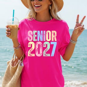 May include: A bright pink short-sleeved t-shirt featuring the text SENIOR 2027 printed in a distressed, multi-colored font. The letters and numbers are colored in shades of yellow, light blue, and pink, creating a vibrant and bold graphic design.
