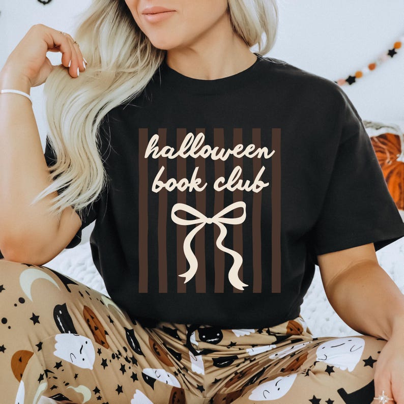 May include: Black t-shirt with the text "halloween book club" in a white, handwritten font. The design includes a cream-colored bow and vertical brown stripes. The shirt is part of a Halloween-themed collection.