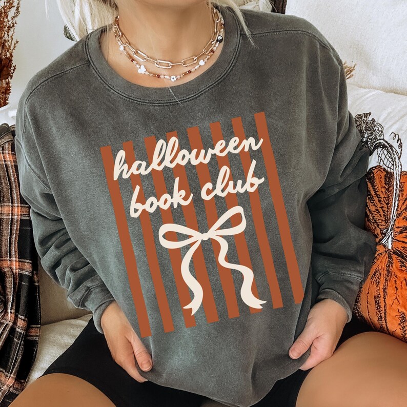 May include: Dark gray sweatshirt with the text "Halloween Book Club" in white script, set against a vertical orange striped background. A white bow graphic is below the text. The sweatshirt is a casual, comfortable style.