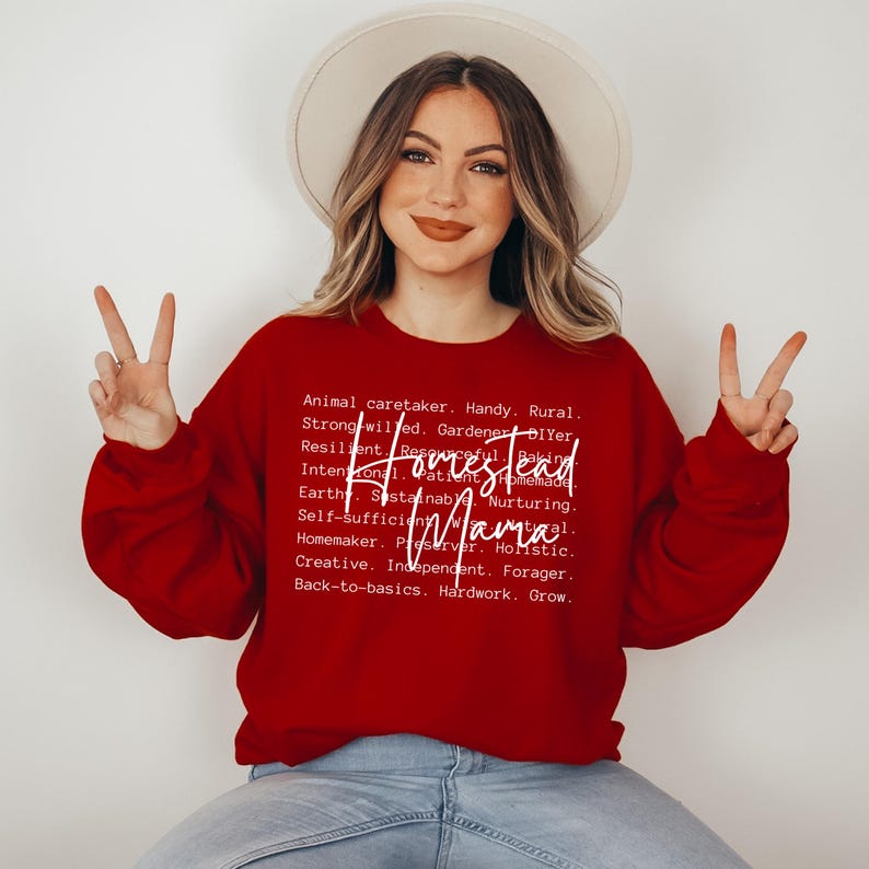 May include: A red sweatshirt with white text that reads "Homestead Mama" and a list of words describing the lifestyle, such as "Animal caretaker", "Handy", "Rural", "Strong-willed", "Gardener", "DIYer", "Resilient", "Resourceful", "Intentional", "Patient", "Homemade", "Earth", "Sustainable", "Nurturing", "Self-sufficient", "Natural", "Homemaker", "Preserver", "Holistic", "Creative", "Independent", "Forager", "Back-to-basics", "Hardwork", "Grow".