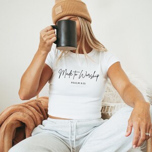 May include: White crop top with the text "Made to Worship" and "Psalm 9:51" in black script. The person is wearing a tan beanie and holding a black mug. The person is sitting down.