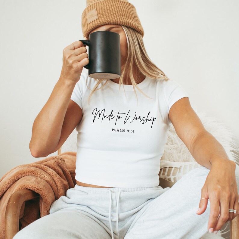 May include: White crop top with the text "Made to Worship" and "Psalm 9:51" in black script. The person is wearing a tan beanie and holding a black mug. The person is sitting down.