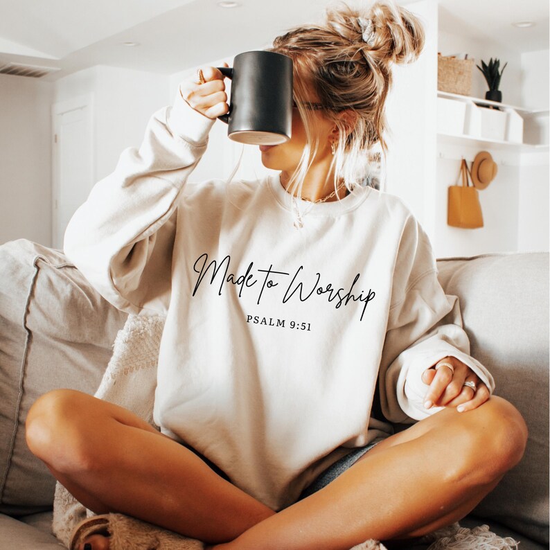 May include: A cream-colored sweatshirt with the text "Made to Worship" and "Psalm 9:51" in black script. A black mug is held up to the face. The sweatshirt is a casual style, suitable for everyday wear.