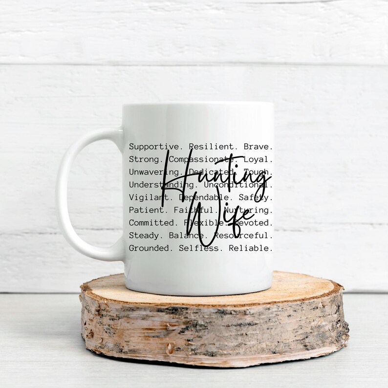 Op de afbeelding: Een witte keramische mok met de woorden "Hunting Wife" in een handgeschreven lettertype. De mok is bedrukt met een lijst met positieve eigenschappen. De mok staat op een houten schijf.