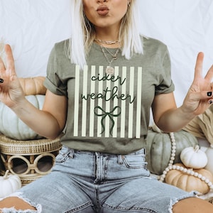 May include: Olive green t-shirt with the words "cider weather" in a script font, set against a striped background with a bow. The model is wearing blue jeans and making a peace sign.