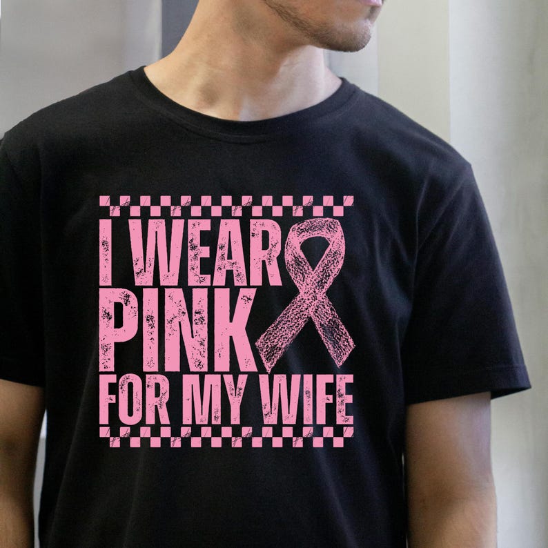 May include: Black t-shirt with the text "I Wear Pink For My Wife" in pink, along with a pink ribbon graphic. The text is arranged in three lines, with a checkered border above and below the text.
