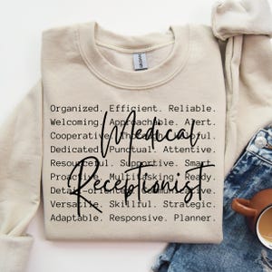 May include: A beige sweatshirt with the text "Organized. Efficient. Reliable. Welcoming. Approachable. Alert. Cooperative. Thoughtful. Dedicated. Punctual. Attentive. Resourceful. Supportive. Smart. Proactive. Multitasking. Ready. Receptionist. Detail-oriented. Communicative. Versatile. Skillful. Strategic. Adaptable. Responsive. Planner." printed in black.