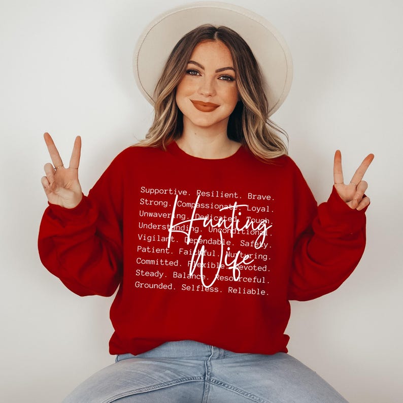 Op de afbeelding: Een rode sweatshirt met de woorden "Hunting Wife" in een cursief lettertype. De sweatshirt heeft ook een lijst met positieve eigenschappen in witte tekst, waaronder "Supportive", "Resilient" en "Brave".