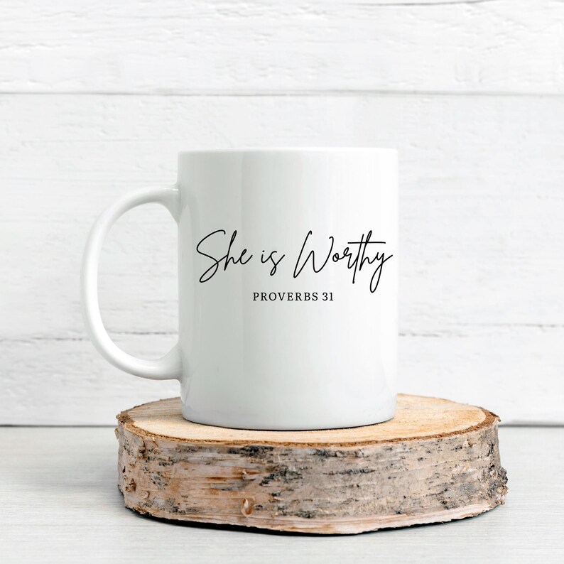 May include: White ceramic mug with the text "She is Worthy" and "Proverbs 31" in black script. The mug is on a round, natural wood slice, set against a white wooden background. A simple and elegant design.
