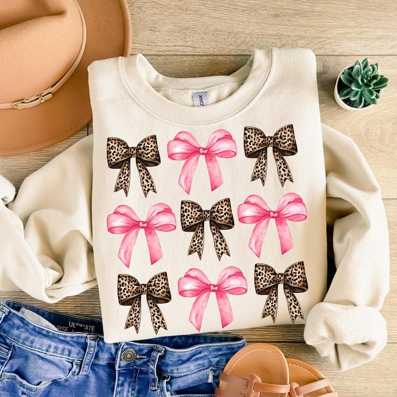 Puede incluir: Una sudadera de cuello redondo color crema con un patr&oacute;n de cuadr&iacute;cula de nueve lazos. El dise&ntilde;o alterna entre lazos de color rosa s&oacute;lido y lazos con estampado de leopardo en tonos marr&oacute;n y negro. La sudadera tiene mangas largas.