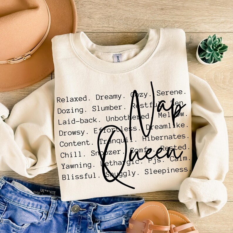 以下が含まれることがあります： ベージュのトレーナーに、黒い文字で「Nap Queen」と書かれています。テキストの周りは、リラックス、夢中、居心地が良い、穏やか、居眠り、睡眠、安らぎ、邪魔されない、努力なし、夢のような、静か、冬眠、休んだ、リラックス、居眠り、快適、あくび、無気力、パジャマ、落ち着く、至福、心地よい、眠気など、昼寝をする感覚を表す言葉が並んでいます。
