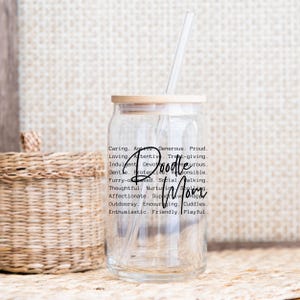May include: A clear glass tumbler with a wooden lid and a straw. The tumbler has a black and white design with the words "Doodle Mom" and a list of adjectives describing a poodle.