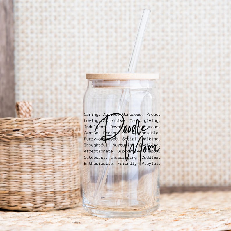 May include: A clear glass tumbler with a wooden lid and a straw. The tumbler has a black and white design with the words "Doodle Mom" and a list of adjectives describing a poodle.