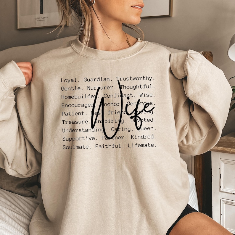 Pu&ograve; includere: Una felpa beige con la parola "Wife" in grandi lettere nere e un elenco di attributi positivi che descrivono una moglie stampati in testo nero.