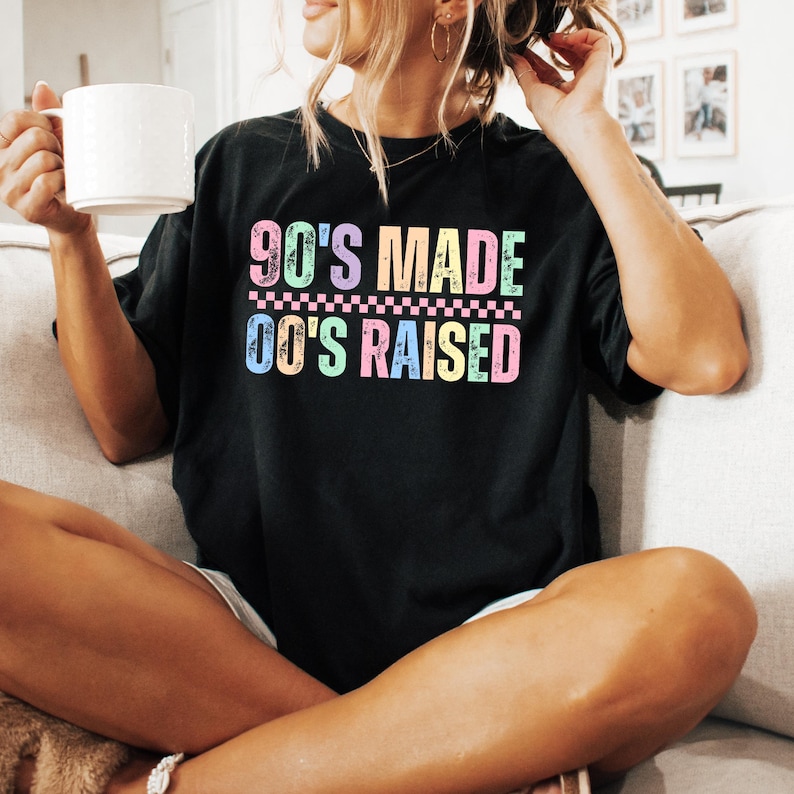 May include: Black t-shirt with the text "90'S MADE 00'S RAISED" in pastel colors. The shirt has a retro design and is likely made of a soft material. The person in the image is holding a white mug.