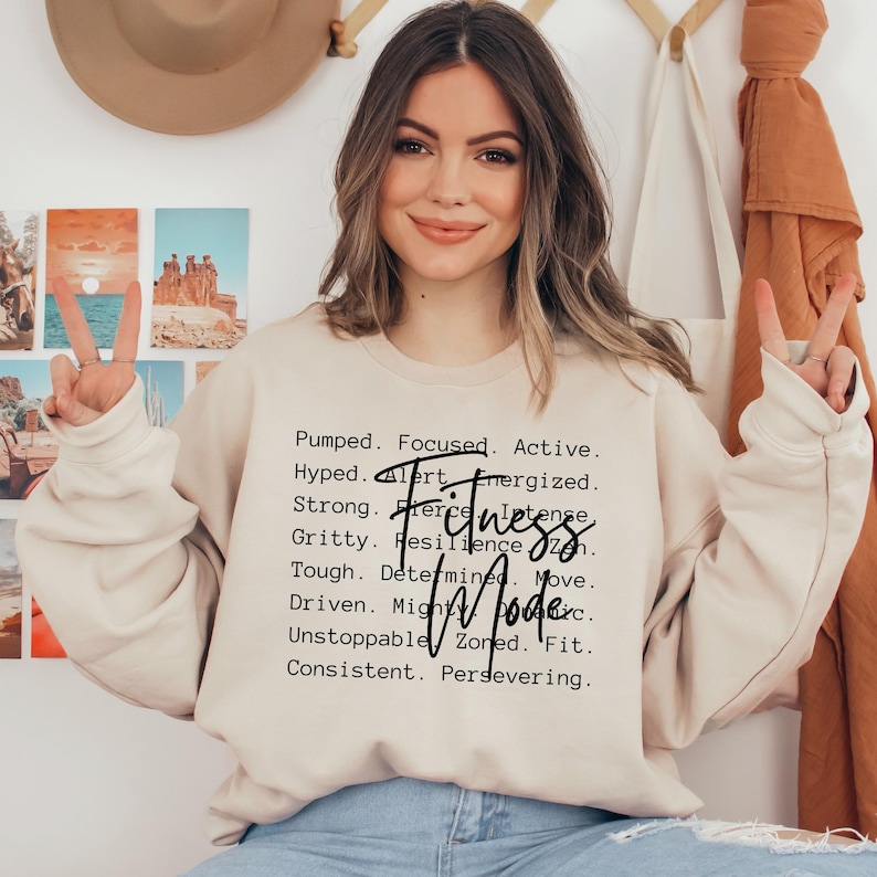 May include: A beige sweatshirt with the words "Fitness Mode" and motivational phrases printed in black. The sweatshirt is worn by a person, and the overall aesthetic is casual and motivational.